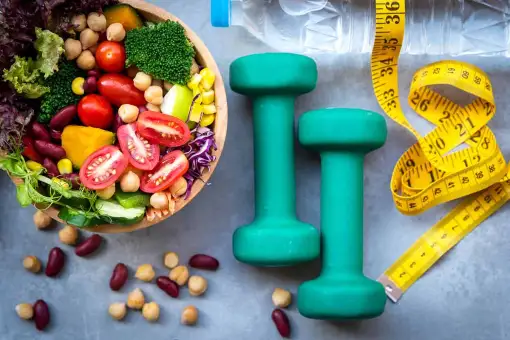 Das Bild zeigt einen ausgewogenen Teller mit Gemüse und Obst, ein Gewicht für das Training und ein Messgerät. Das Bild soll zeigen, dass diese 3 Komponenten grundlegend für ein gesundes, ausgewogenes Leben sind.